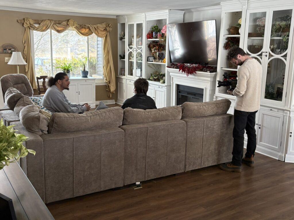A man and a teen boy sitting on a couch in the living room, talking while being filmed by a cameraman.